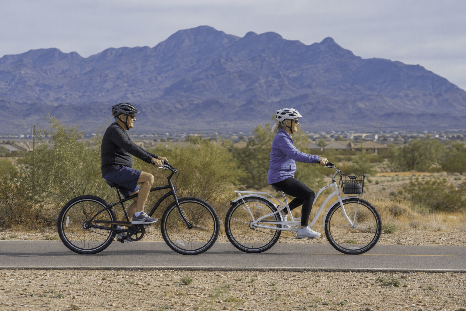 couple in their late 60s, different genders, riding cruiser-style bicycles side by side along a paved multi-use path in southern Goodyear, Arizona